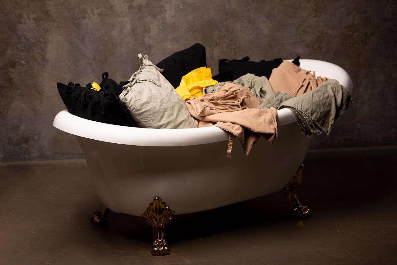 A vintage clawfoot bathtub filled with assorted clothes against a textured wall.